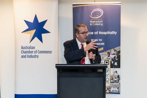 John Hart OAM speaks at the Welcome Back to Parliament event, standing at a podium between banners for the Australian Chamber of Commerce and Industry and Restaurant & Catering Australia.