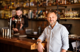 Owner of a café standing confidently at the bar while a staff member prepares drinks in the background — representing small business operators supported by R&CA’s IR service.