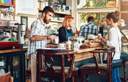 A barista and staff serve customers inside a busy café, with coffee machines, pastries, and food items displayed on the counter, reflecting a typical Australian hospitality setting focused on customer service and food safety.