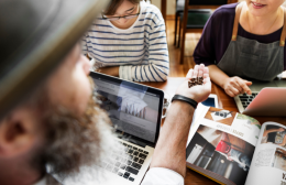 A café owner and team discuss marketing strategies around a table with laptops, coffee, and printed materials, highlighting collaboration and planning in a foodservice business.