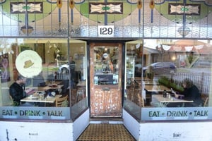 Street view of a café with stained glass windows and the number 129 above the entrance. The front glass panels display the words “EAT • DRINK • TALK,” with two people seated inside at tables. The café has a rustic wooden door and warm interior lighting, giving it a welcoming, local atmosphere.