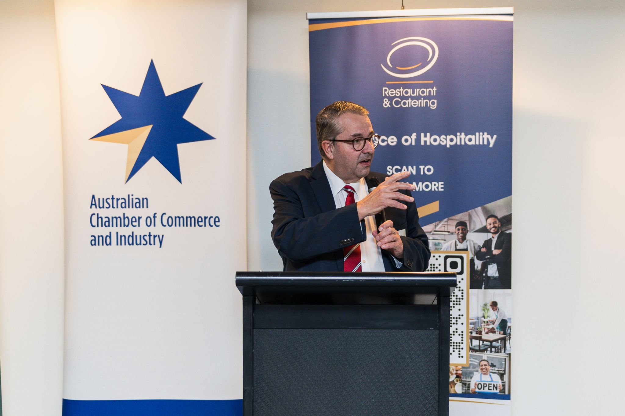 John Hart OAM speaks at the Welcome Back to Parliament event, standing at a podium between banners for the Australian Chamber of Commerce and Industry and Restaurant & Catering Australia.