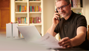 Middle-aged man reviewing documents while speaking on the phone at a home office desk, with a laptop and files in front of him.