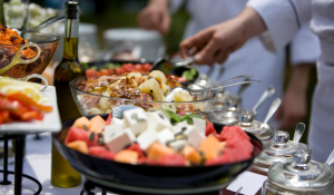 Chefs serving fresh catering dishes outdoors, with a colourful spread including watermelon, feta, and roasted vegetables.