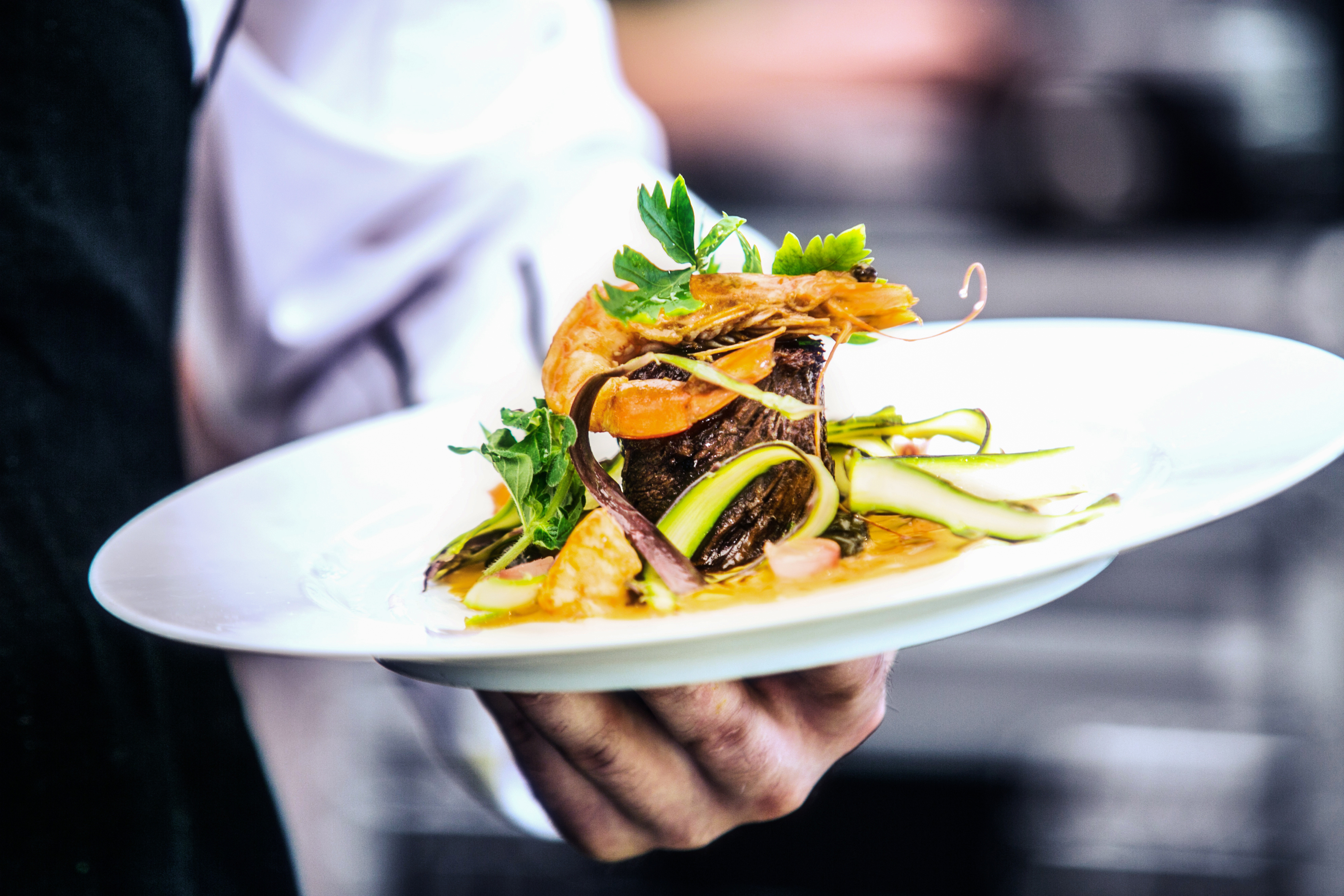 Chef holding a white plate with a plated dish of braised beef, king prawns, zucchini ribbons, and fresh herbs in a restaurant kitchen setting.