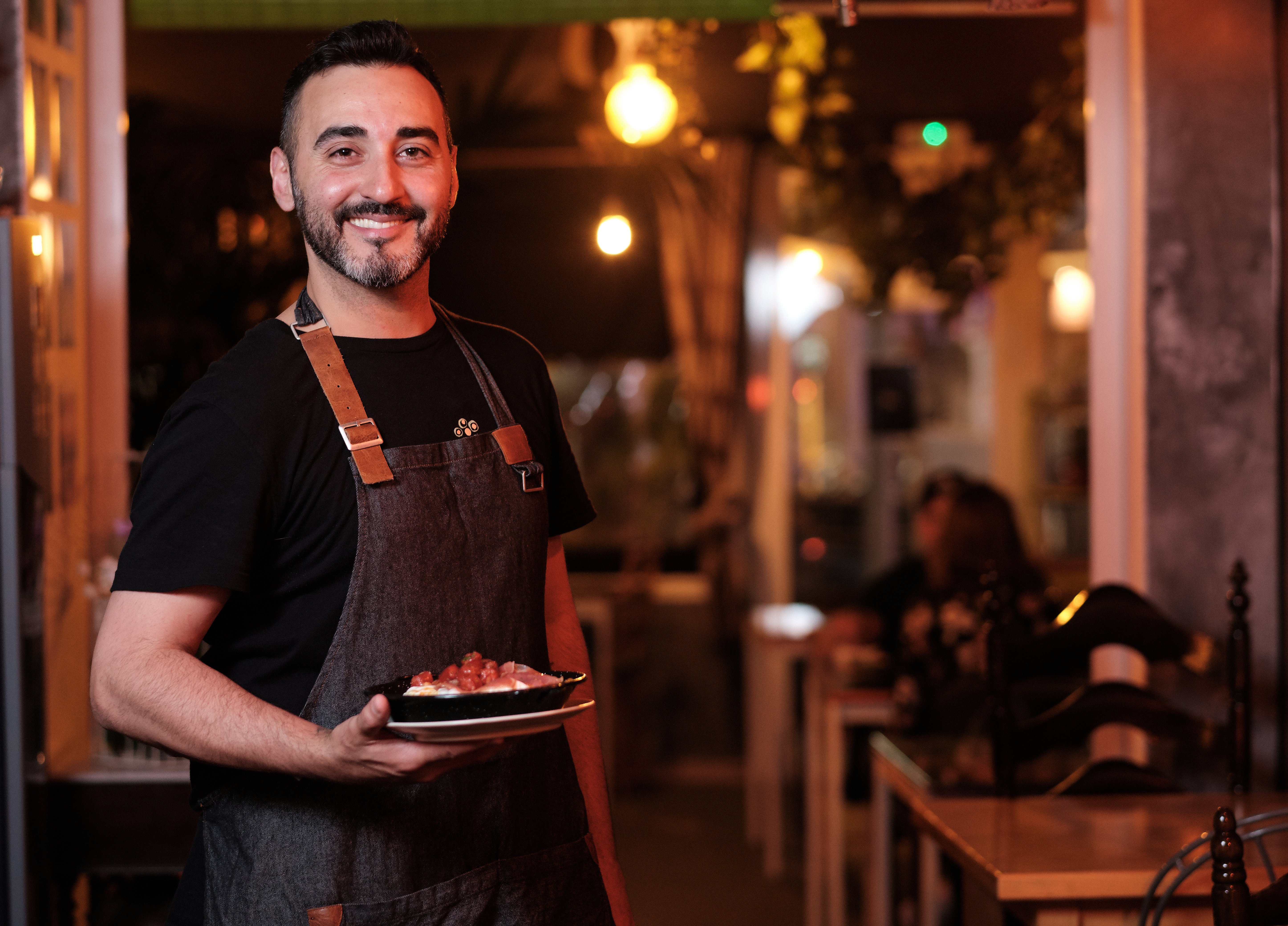 Smiling restaurant staff member in an apron holding a plated dish, standing in a warmly lit dining space with guests in the background.