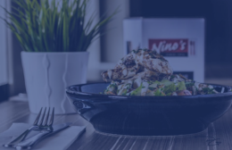 Grilled chicken salad served in a black bowl on a wooden table at Nino’s Restaurant, with a white plant pot and Nino’s branded menu in the background.