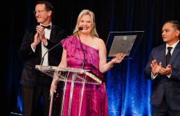 A woman in a bright pink dress stands at a podium holding an award, smiling as two men applaud beside her during a hospitality awards ceremony.