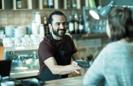 Barista in a café smiling as he processes a digital payment for a customer, representing how technology enhances everyday hospitality service.