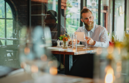 A man dining alone at a restaurant table, relaxed and reading his phone over a glass of wine.