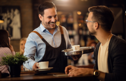 A smiling waiter serves coffee to a regular customer at the bar of a warmly lit cafe