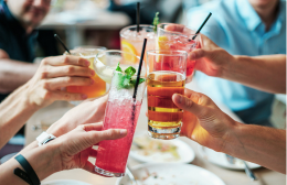People raising a mix of colourful drinks in a cheers gesture, including mocktails and non alcoholic beverages, at a dining table.