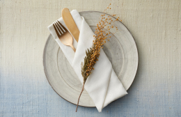 A neutral table setting with a ceramic plate, white folded napkin, gold cutlery and a small sprig of dried native foliage.