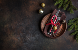 Rustic Christmas table setting with cutlery tied in a red ribbon on a wooden plate, styled with greenery on a dark background.