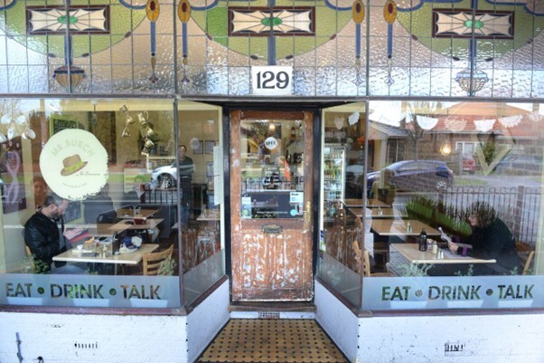 Street view of a café with stained glass windows and the number 129 above the entrance. The front glass panels display the words “EAT • DRINK • TALK,” with two people seated inside at tables. The café has a rustic wooden door and warm interior lighting, giving it a welcoming, local atmosphere.